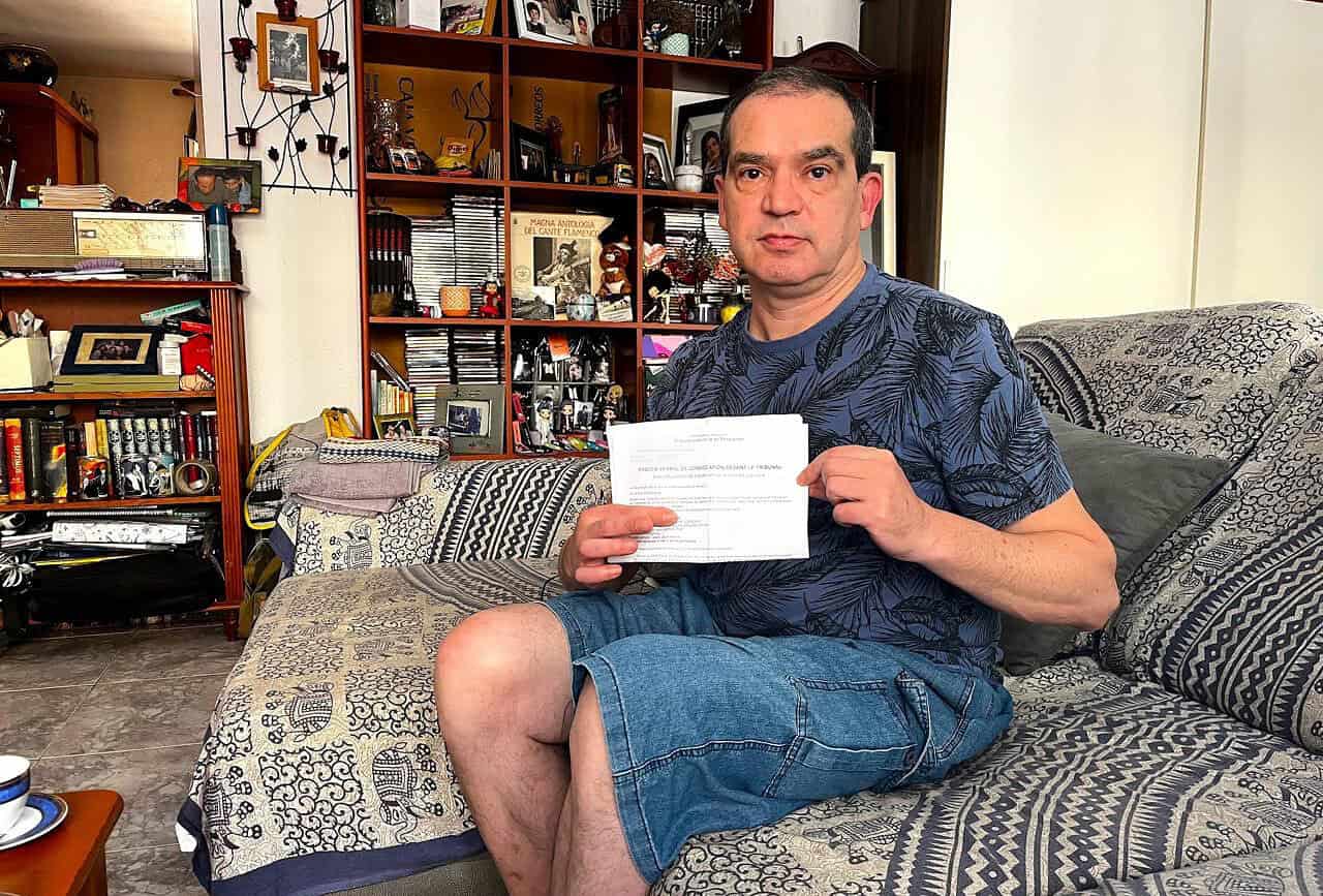 Homme assis sur un canapé, tenant un document, dans un intérieur avec étagères remplies de livres et objets, illustrant une situation liée à un contexte de trafic de migrants.