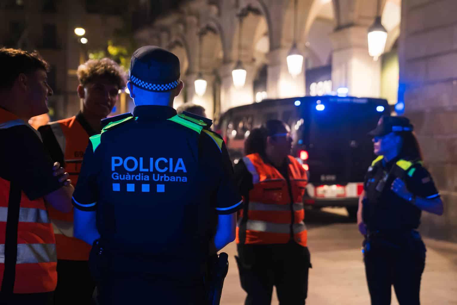 Policier de la Guardia Urbana en discussion avec des agents de sécurité lors d'une intervention nocturne dans une rue éclairée.