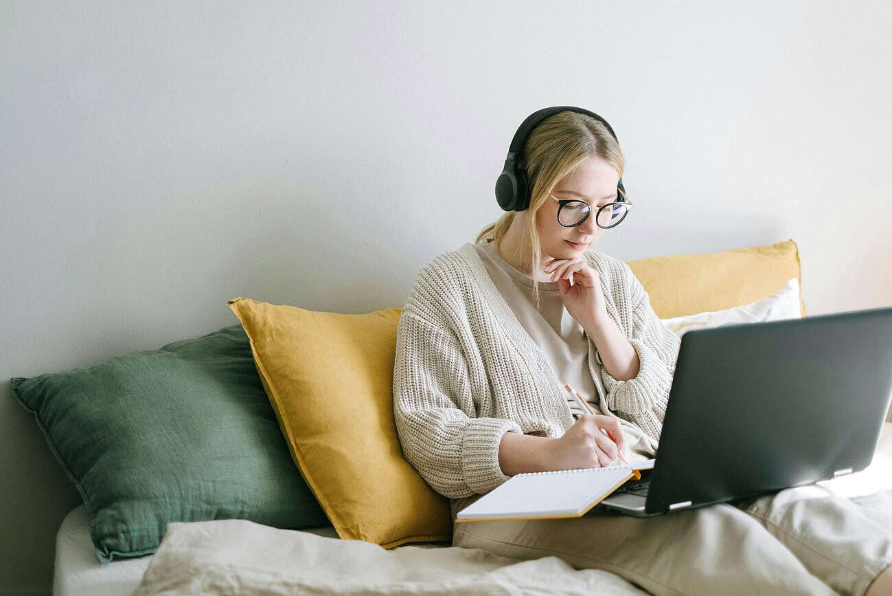 Femme détendue portant des écouteurs, travaillant ou se relaxant avec un ordinateur portable et un carnet, dans un cadre confortable à la maison.