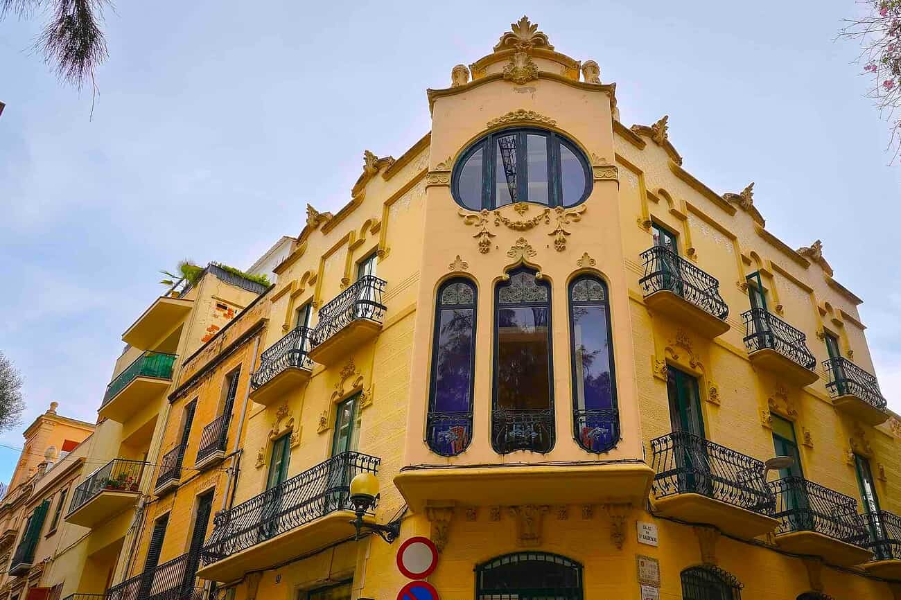 Balconnières et façade colorée d’un bâtiment moderniste à Sitges, sous un ciel clair, illustrant le charme architectural de cette ville méditerranéenne proche de Barcelone.
