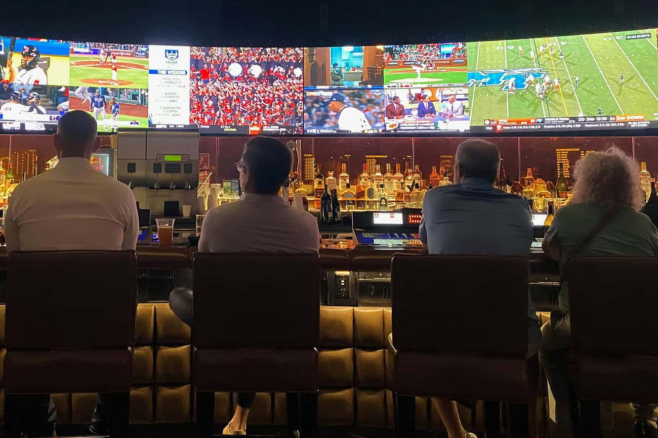 La grippe footballistique : quand les Espagnols sèchent le bureau pour le Mondial 2 Quatre personnes regardent un grand écran de télévision diffusant un match de football dans un bar, avec plusieurs autres écrans en arrière-plan montrant différents événements sportifs.