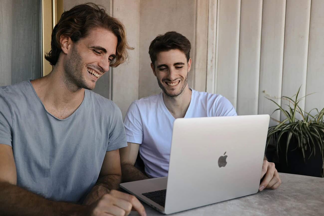 Barcelone : une ville qui travaille avec le sourire 2 Balcon avec deux jeunes hommes souriants travaillant ensemble sur un ordinateur portable à Barcelone, illustrant une atmosphère de travail positive et conviviale dans la ville.