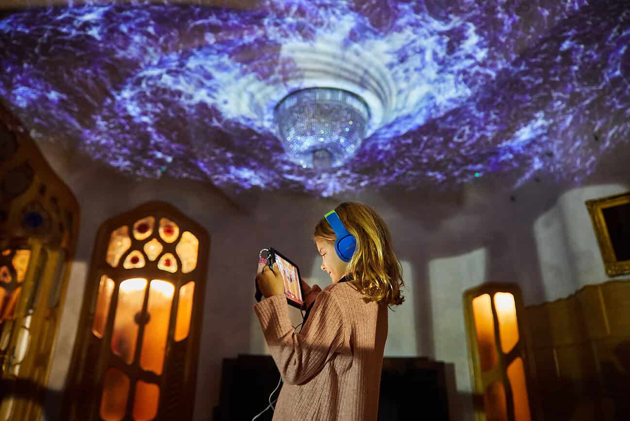 Casa Batlló : l’univers féerique de Gaudí s’illumine avec “Nuit d’Hiver” 10 Une jeune fille écoute de la musique dans une salle de Casa Batlló, illuminée par un spectacle lumineux féerique projeté sur le plafond, créant une ambiance magique et immersive.