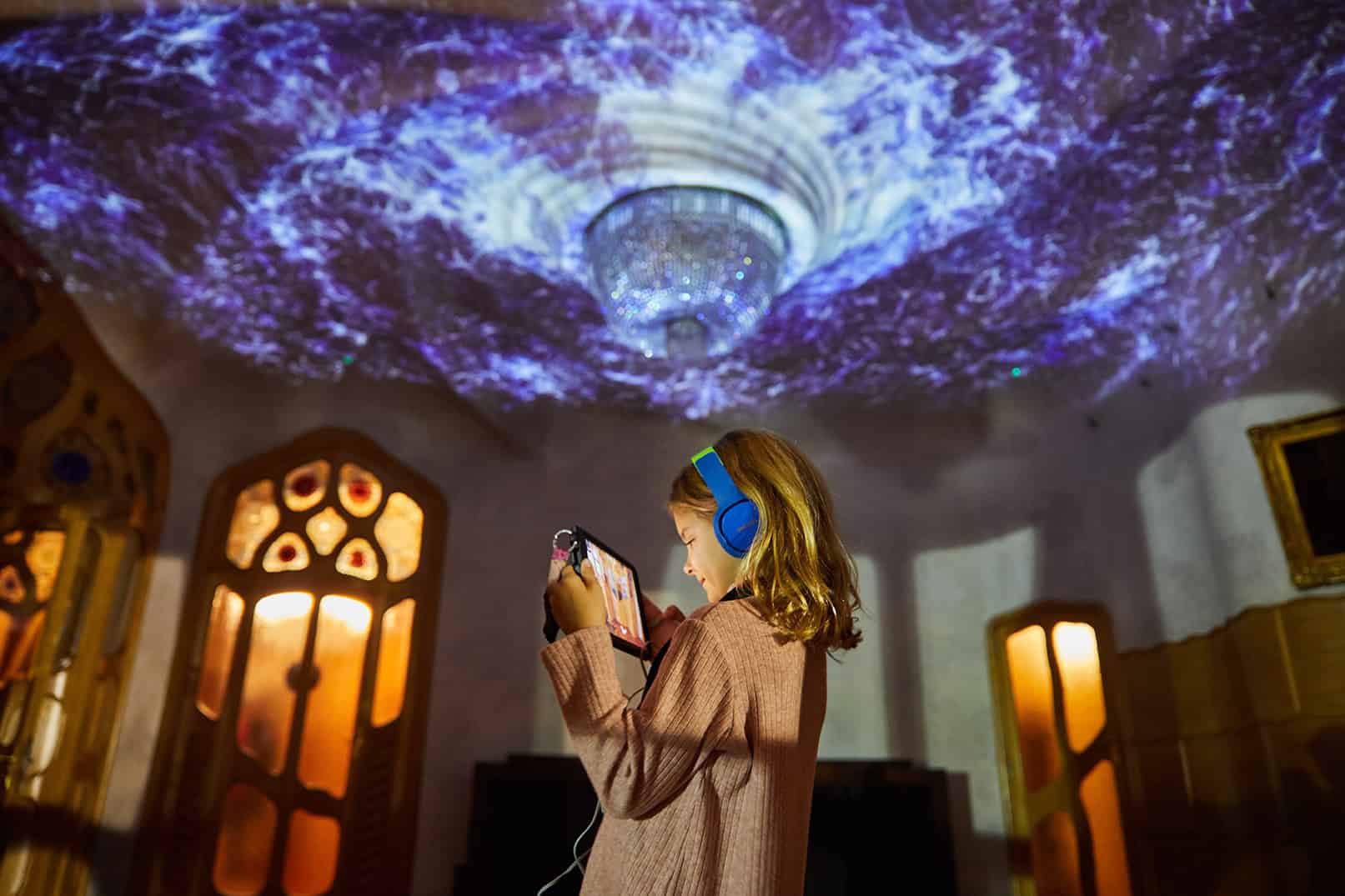 Une jeune fille écoute de la musique dans une salle de Casa Batlló, illuminée par un spectacle lumineux féerique projeté sur le plafond, créant une ambiance magique et immersive.