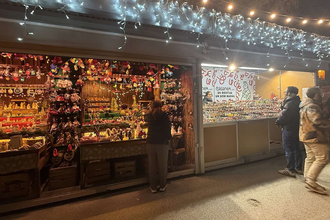 5 cadeaux catalans pour faire douter votre famille de votre santé mentale 2 Vente de décorations de Noël colorées et festives dans un marché de Noël à Barcelone, avec des visiteurs qui découvrent des objets artisanaux et des souvenirs saisonniers.