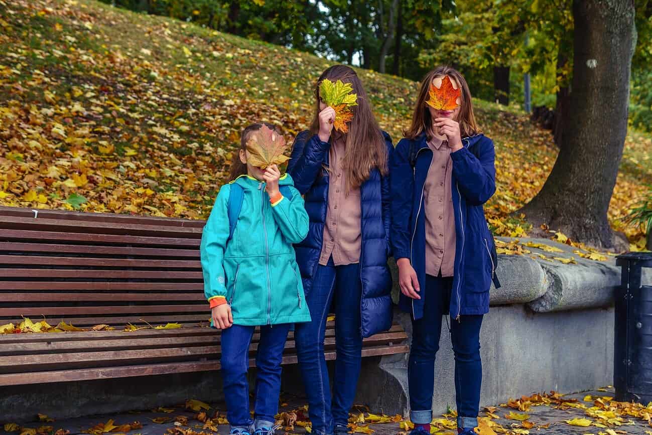 Pourquoi les Espagnols font des blagues le 28 décembre 1 Trois jeunes filles dans un parc automnal, portant des manteaux, avec des feuilles d'automne sur le visage, illustrant la tradition humoristique espagnole du 28 décembre.