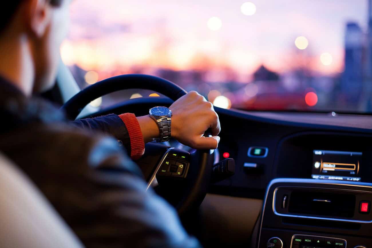 Conducteur au volant d'une voiture, mains sur le volant, avec un paysage urbain flou en arrière-plan au coucher du soleil.