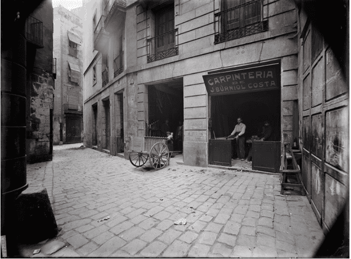 Vue d'une vieille rue commerçante à Barcelone, avec un artisan travaillant dans une boutique de menuiserie, pavés traditionnels et bâtiments anciens.