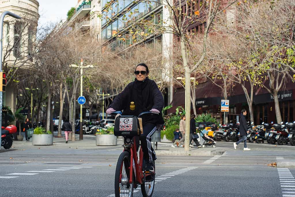 Un homme souriant portant des lunettes, pédalant à vélo dans une rue urbaine animée, entouré d'arbres et de bâtiments modernes, illustrant un mode de vie actif et écologique.