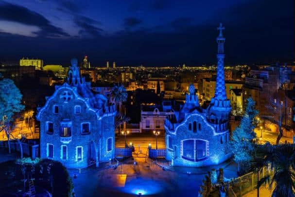 Le Park Güell de Barcelone éclairé en bleu, mettant en valeur ses structures architecturales uniques et colorées, avec la ville en arrière-plan sous un ciel nocturne.