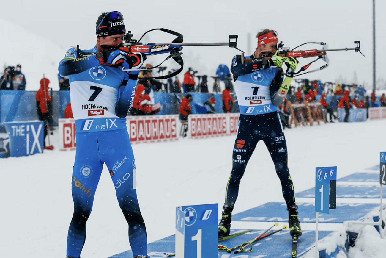 Comment le système de tir a évolué dans le biathlon moderne 4 Deux biathlètes en position de tir lors d'une compétition de biathlon, avec un public en arrière-plan et un parcours enneigé.