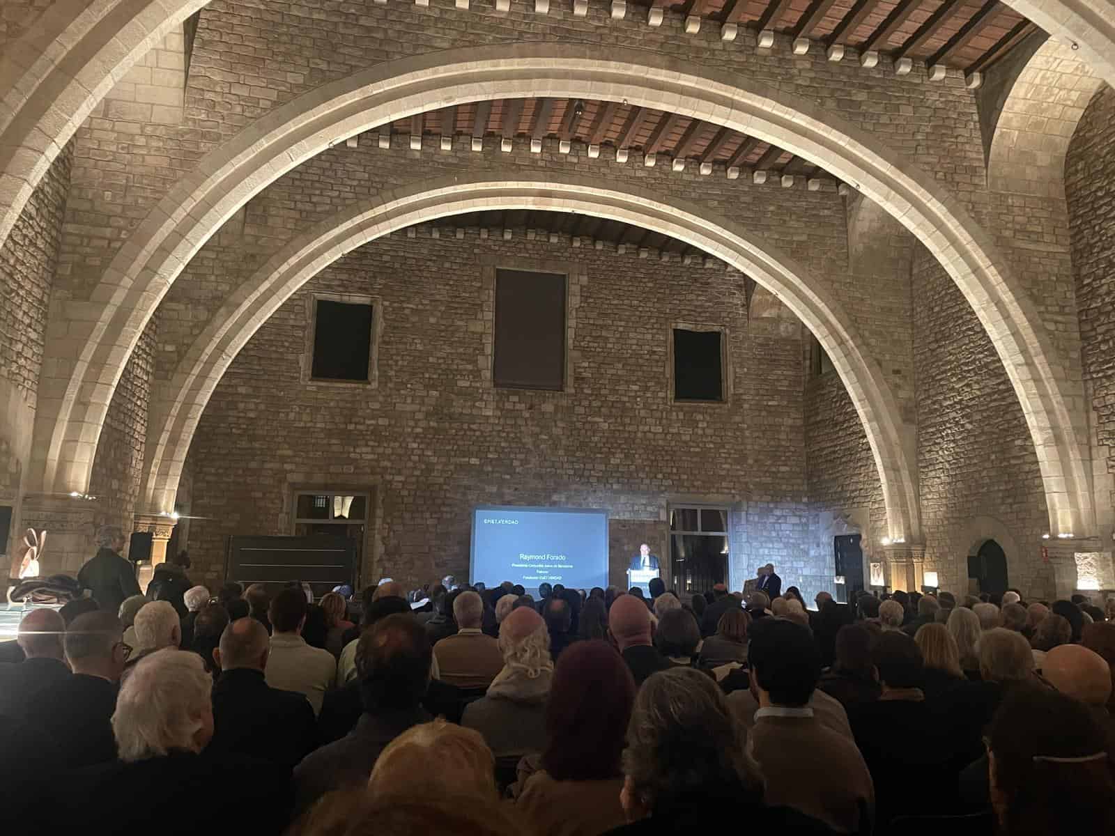 Valls en tête d'une conférence à Barcelone pour lutter contre l'antisémitisme, entouré d'un public nombreux dans une salle historique avec des arches en pierre.