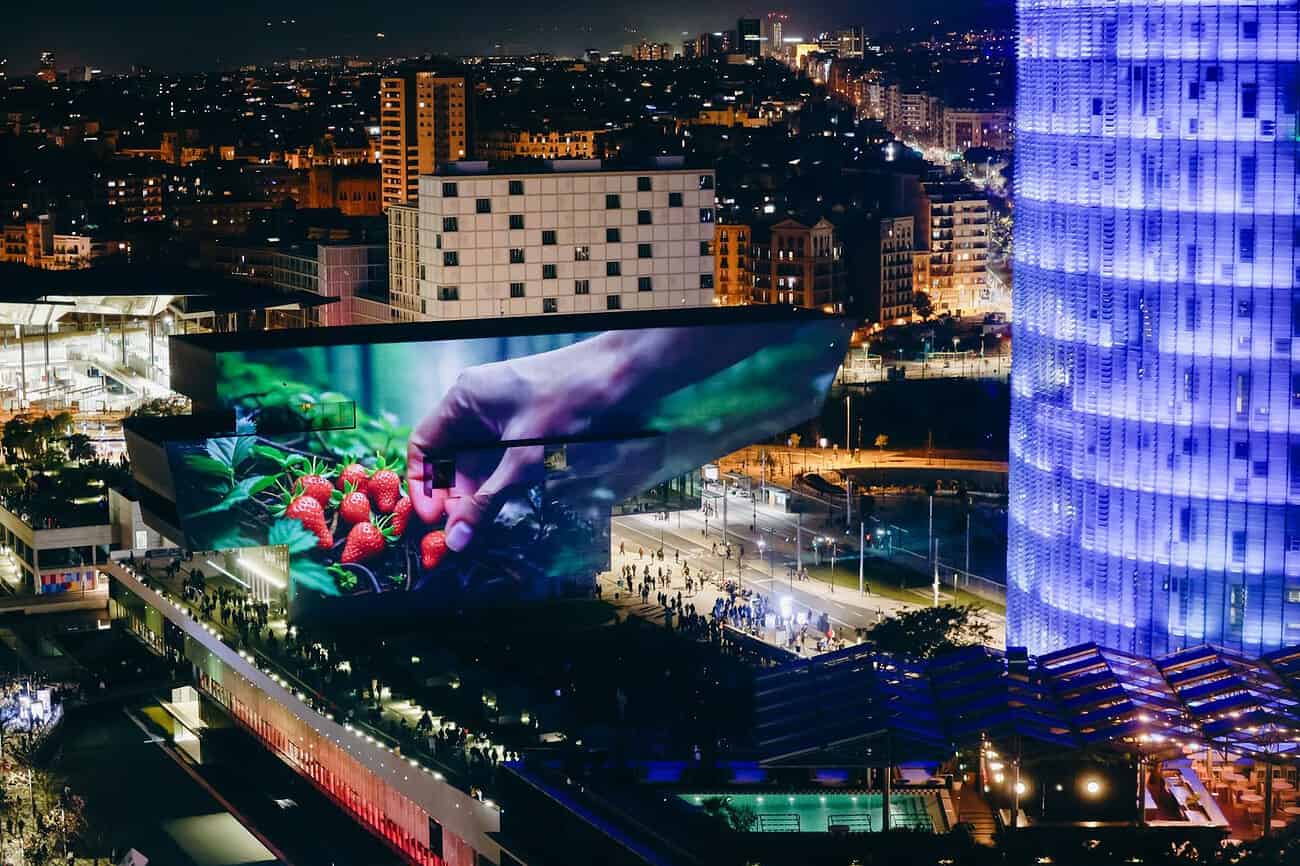 Que faire à Barcelone le week-end du 6 au 8 février 1 Une grande vitrine numérique illuminée dans le centre-ville de Barcelone, affichant des images colorées de fruits et de mains manipulant des produits frais, avec la ville en arrière-plan la nuit.
