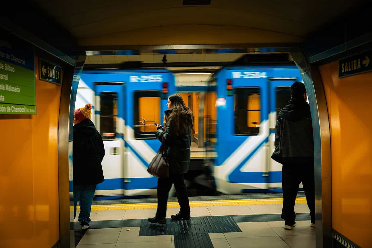 Trois personnes attendent sur le quai du métro dans une station souterraine, prêtes à monter dans le train bleu qui arrive.