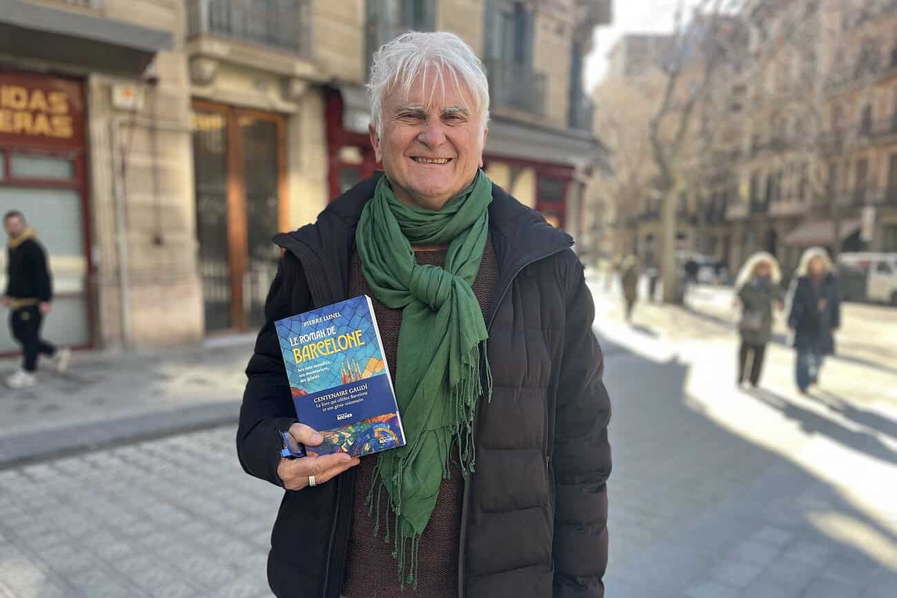 Livre "Le Roman de Barcelone" présenté par un homme souriant dans une rue de Barcelone, célébrant le centenaire de Gaudí avec un livre en main.
