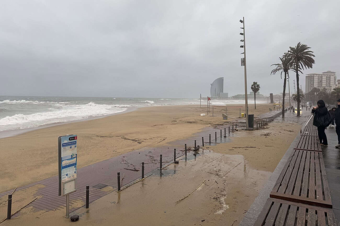 Balade pluvieuse sur la plage de Barcelone avec vue sur le bâtiment W Barcelona, palmiers et promenade en bois, ambiance nuageuse et tempétueuse.