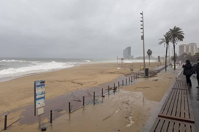 Balade pluvieuse sur la plage de Barcelone avec vue sur le bâtiment W Barcelona, palmiers et promenade en bois, ambiance nuageuse et tempétueuse.