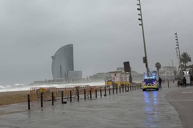 Plage de Barcelone par temps pluvieux avec des vagues, un ciel nuageux et une promenade vide, mettant en valeur l'atmosphère humide et venteuse de la côte méditerranéenne.
