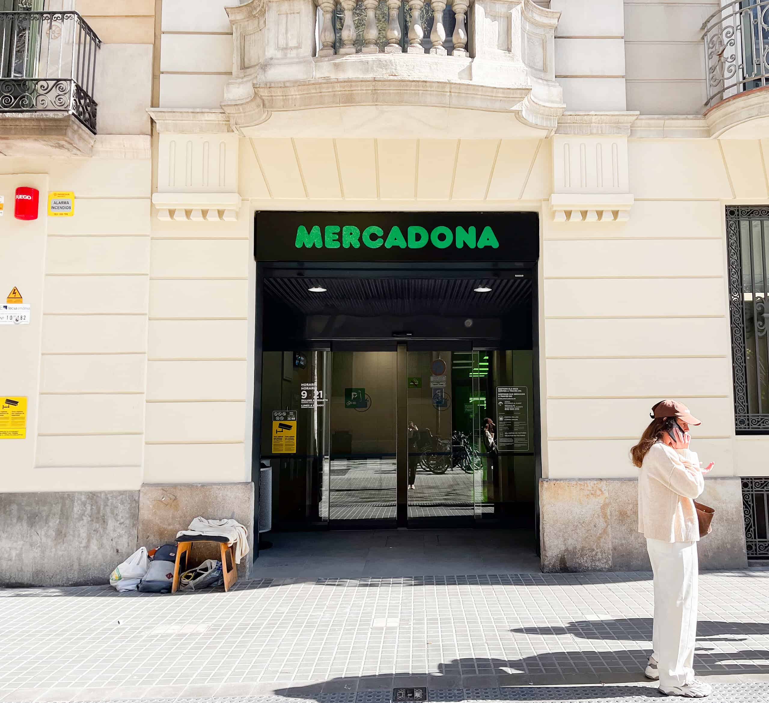 Balayage de l'entrée du supermarché Mercadona à Madrid, avec une femme portant un masque et un sac à la main, sous un bâtiment historique.