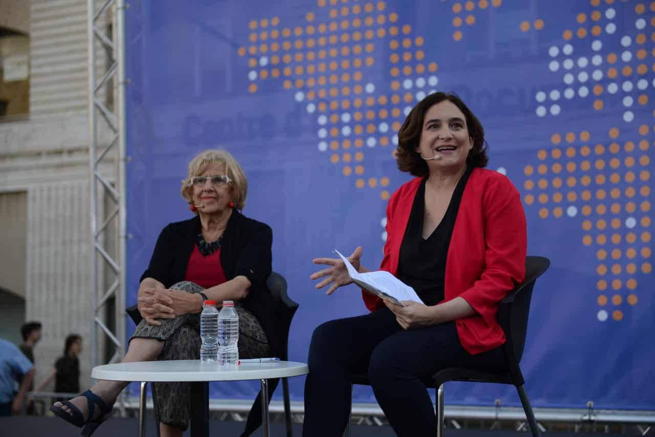 Deux femmes en discussion lors d'une conférence sur le municipalisme, avec un fond illustrant une carte du monde en points colorés, évoquant les expériences de Barcelone et Madrid.