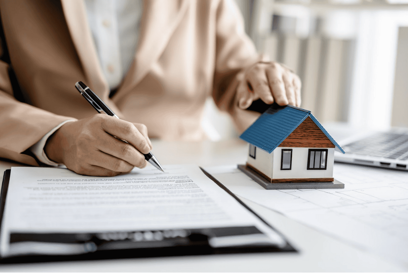 Personne signant un contrat immobilier avec une petite maison en modèle réduit sur la table, illustrant le marché immobilier à Barcelone.