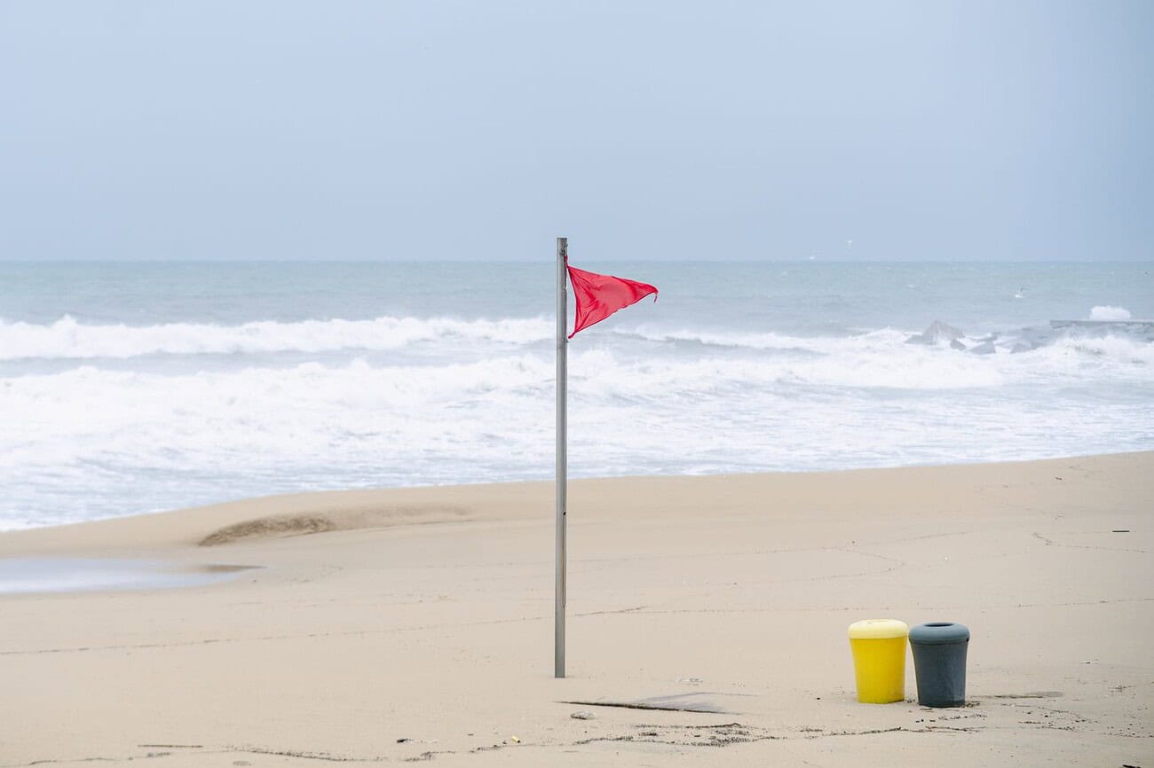 Une violente tempête attendue à Barcelone 1 Plage de Barcelone avec drapeau rouge, symbolisant la sécurité et la vigilance en bord de mer, sous un ciel nuageux, avec deux poubelles jaunes et noires sur le sable.