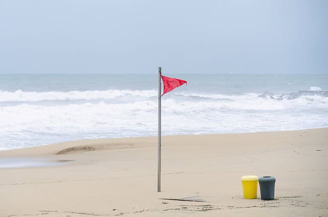 Equinox : toute l'actu de Barcelone et les bons plans de la ville 9 Plage de Barcelone avec drapeau rouge, symbolisant la sécurité et la vigilance en bord de mer, sous un ciel nuageux, avec deux poubelles jaunes et noires sur le sable.