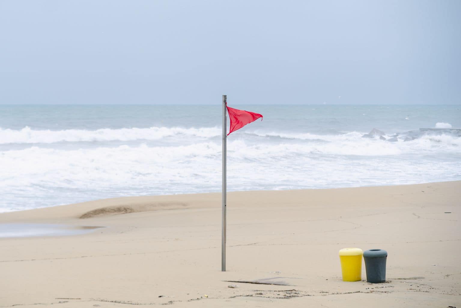Plage de Barcelone avec drapeau rouge, symbolisant la sécurité et la vigilance en bord de mer, sous un ciel nuageux, avec deux poubelles jaunes et noires sur le sable.
