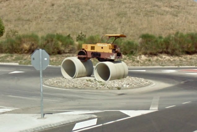 Vue d'un rond-point atypique avec un engin de construction sur des tuyaux en béton, situé dans un environnement rural, illustrant une installation créative ou humoristique.