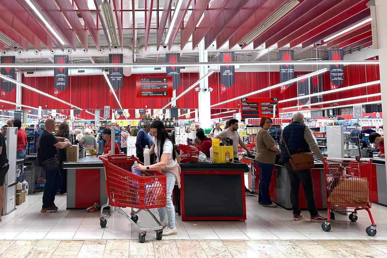 Clients faisant leurs courses dans un supermarché à la frontière franco-espagnole, avec caddies et caisses enregistreuses visibles.